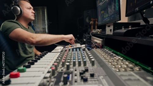 a focused male sound engineer wearing headphones sits at a mixing desk recording new track photo