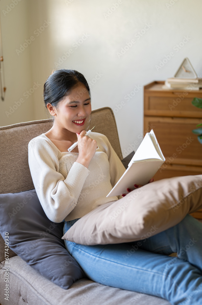 An attractive Asian woman is reading a book or keeping diary while relaxing in her living room. An attractive Asian woman is reading a book or keeping diary while relaxing in her living room.
