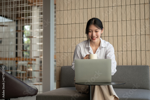 A charming Asian businesswoman is working remotely at a cafe , using her laptop and sipping coffee.