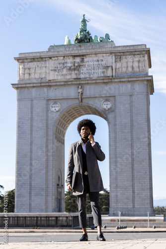 Vertical photo of a young latin businessman in front of the victory arch in Madrid while talking on his mobile phone and holding his laptop under his arm. © Martin