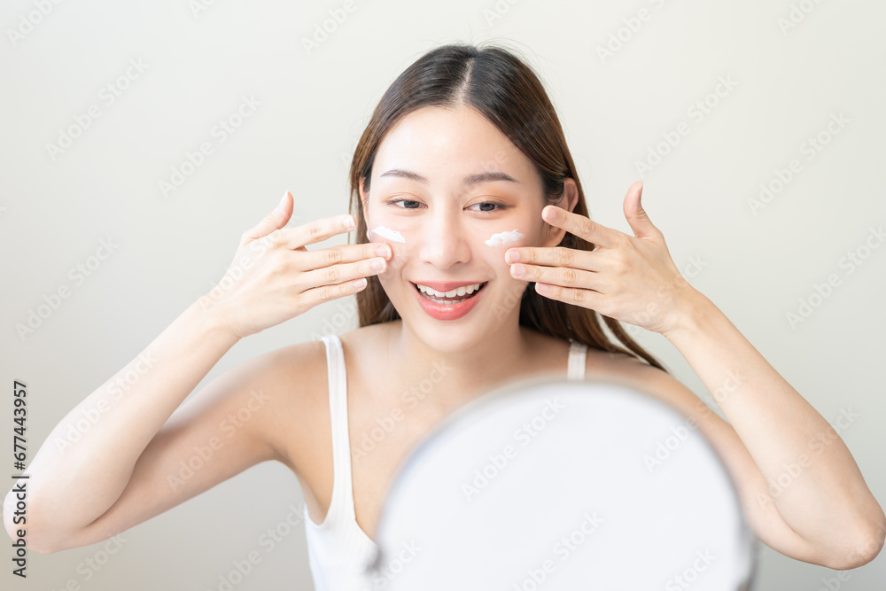 Fresh healthy skin, beautiful smile of asian young woman, girl looking at mirror, applying moisturizer on her face, putting cream treatment before makeup cosmetic routine at home. Facial Beauty. Fresh healthy skin, beautiful smile of asian young woman, girl looking at mirror, applying moisturizer on her face, putting cream treatment before makeup cosmetic routine at home. Facial Beauty.