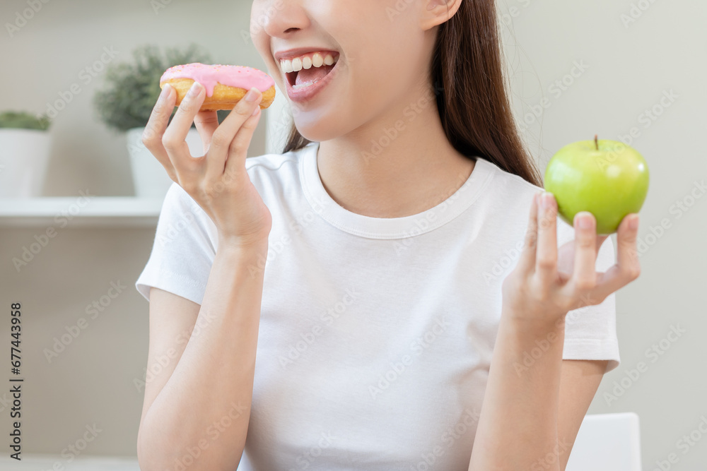Diet, Dieting. Close up hand of asian young woman confused with food temptation choose, choice pink sweet donut or green apple at home, eat food for good healthy, health when hungry, weight loss. Diet, Dieting. Close up hand of asian young woman confused with food temptation choose, choice pink sweet donut or green apple at home, eat food for good healthy, health when hungry, weight loss.