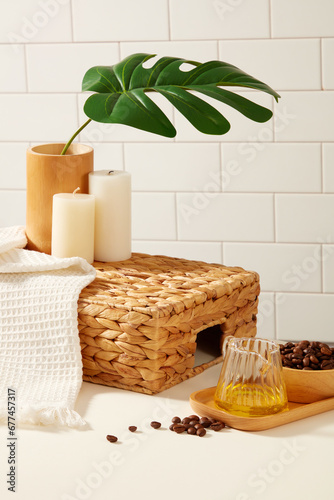 On the white tile background, coffee beans on wooden tray decorated with towel, candle and green monstera leaves. Blank space on basket for display bath products