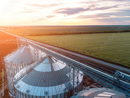 Grain silos on a green field background with warm sunset light. Grain elevator. Metal grain elevator in agricultural zone. Agriculture storage for harvest. Aerial view of agricultural factory. Nobody.