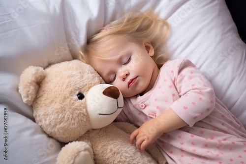 Little Caucasian girl with dark hair in shirt sleeps sweetly in company of best friend teddy bear seeing pleasant dreams. Little girl has sweet dreams in bed with favorite toy small teddy bear