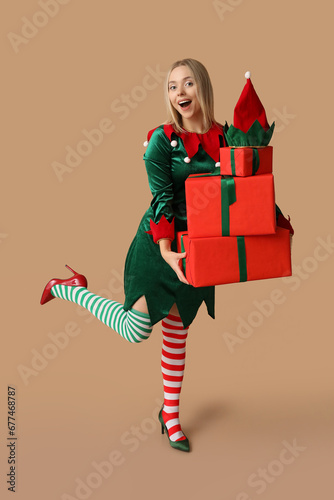 Young woman in elf costume holding Christmas gift boxes on brown background