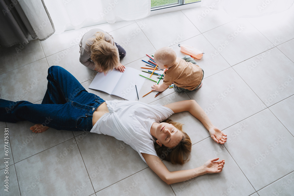 Tired mother lying on floor while her children drawing nearby. - obrazy, fototapety, plakaty 