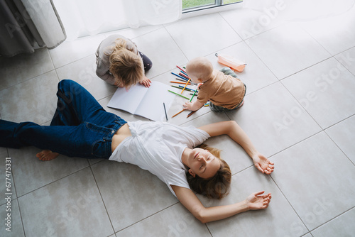 Tired mother lying on floor while her children drawing nearby.