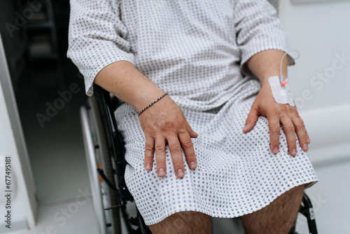 Close up of man in wheelchair with IV cannula in hand. Overweight patient in hospital gown waiting for medical examination in hospital.