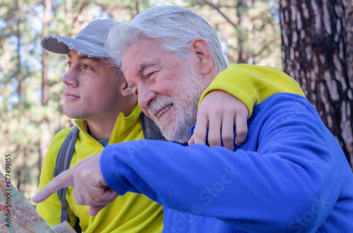 Fotografija Young grandson and old grandfather consulting trail map while hiking in mountain forest enjoying nature and healthy activity