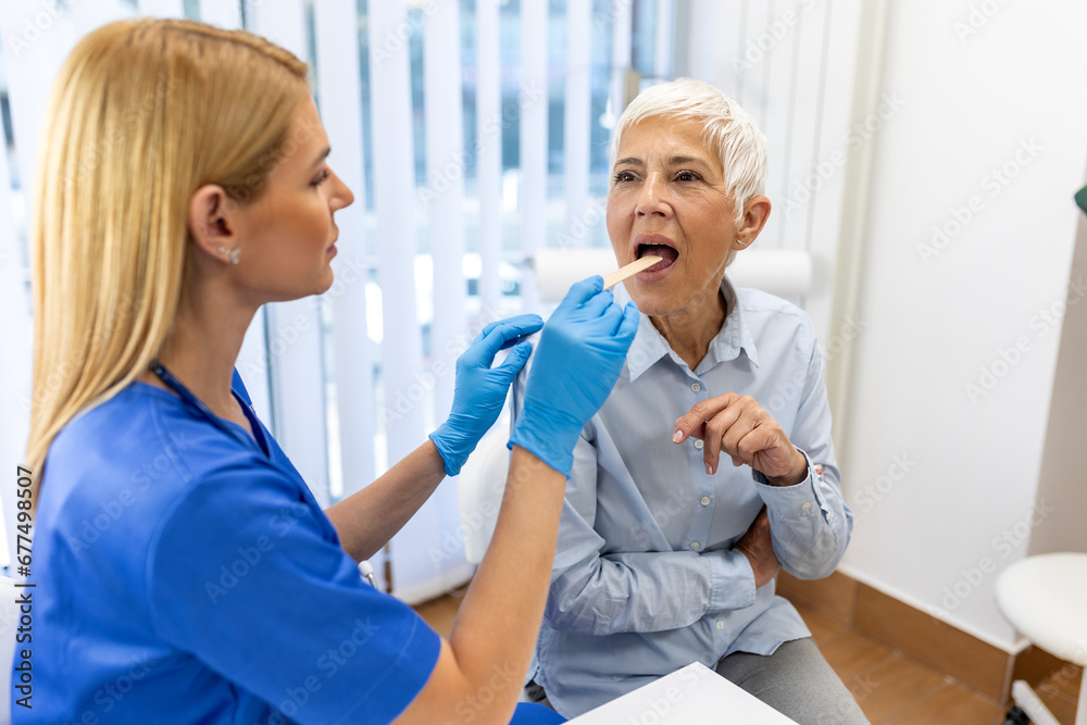 Obraz premium Senior patient opening her mouth for the doctor to look in her throat. Female doctor examining sore throat of patient in clinic. Otolaryngologist examines sore throat of patient.