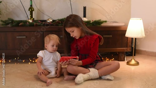 Two small girls sit and watch video on phone. Children busy with gadget. Time for happy Christmas. Children together joy in home.