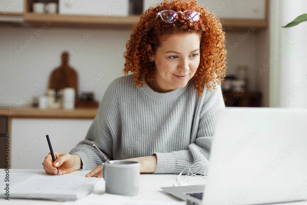Portrait of happy female student watching webinar or tutorial studying online on laptop sitting at kitchen table, noting down after teacher or lecturer, looking at screen with interest - obrazy, fototapety, plakaty 