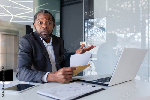 Adult man surprised holding open envelope in hands, shocked by news, disappointing news, african american boss sitting in bright office looking at camera and spreading hands.