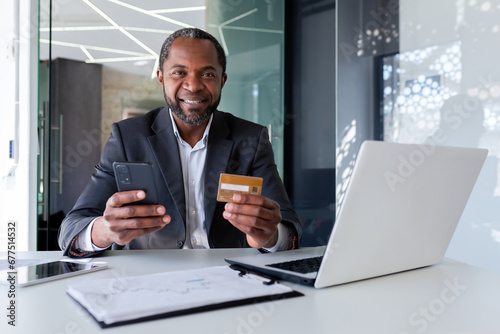 Senior African American man in suit sitting at desk in office. He is holding a credit card and a phone. Successfully paid online purchases, orders, bills. Happy looking at camera.
