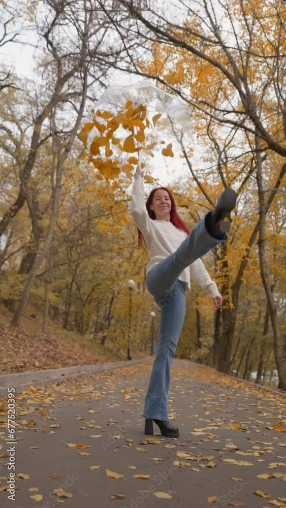 Happy Woman in Jeans and a White Sweater Walks and Lifts her Leg into Splits and Raises an Umbrella from Which Autumn Leaves are Flying. Slow Motion, Vertical Video. Happy Woman in Jeans and a White Sweater Walks and Lifts her Leg into Splits and Raises an Umbrella from Which Autumn Leaves are Flying. Slow Motion, Vertical Video.