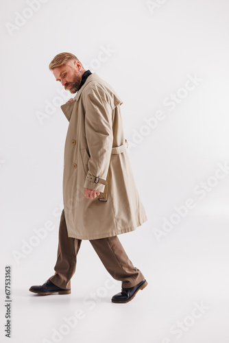 Trendy blonde man in classic trench coat walking on grey 