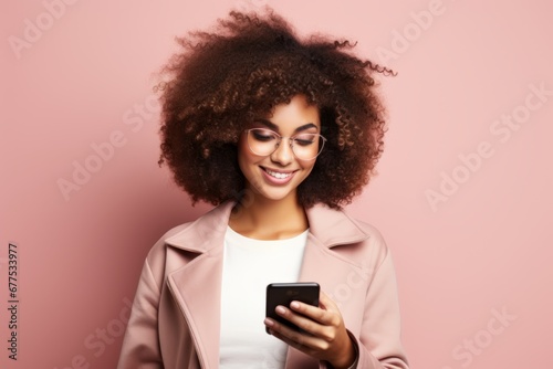Portrait of beautiful positive girl with Afro haircut in pink coat typing text message on smartphone, enjoying online communication, using app, chatting. Isolated on pink studio background.