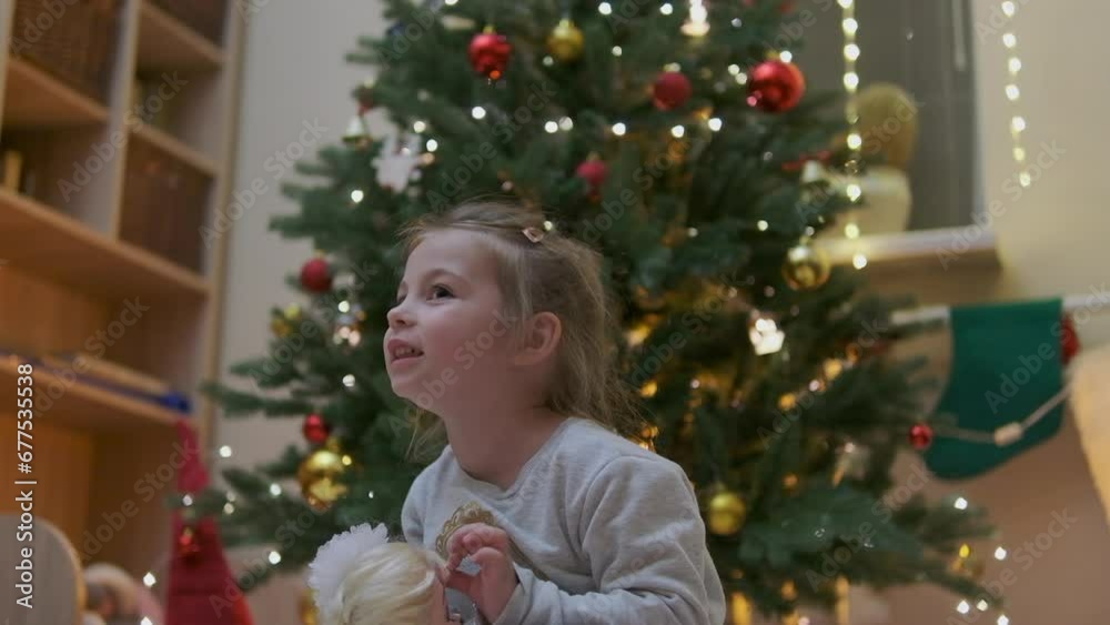 Little beautiful girl near the Christmas tree. Girl with a doll are playing Christmas. Beautiful girl with long hair Little beautiful girl near the Christmas tree. Girl with a doll are playing Christmas. Beautiful girl with long hair