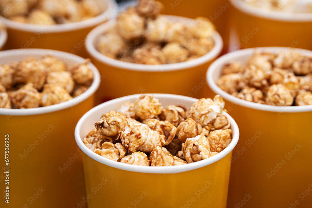 delicious sweet caramel brown popcorn. in a yellow orange glass close-up. boxes of popcorn on sale on the counter in the store. Paper bucket with popcorn, concept of watching TV or cinema. delicious sweet caramel brown popcorn. in a yellow orange glass close-up. boxes of popcorn on sale on the counter in the store. Paper bucket with popcorn, concept of watching TV or cinema.