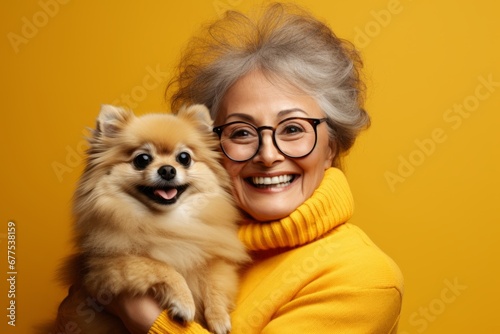 Portrait of smiling Caucasian senior woman in yellow sweater posing with cute Pomeranian spitz puppy. Happy lady hugs her beloved pet. Love between human and dog. Yellow studio background, copy space. © Georgii