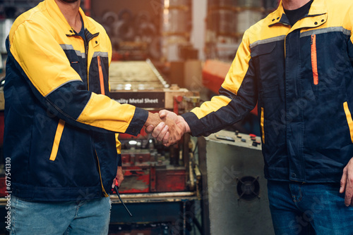 Engineer and industrial worker in uniform shaking hands in large metal factory hall and talking.