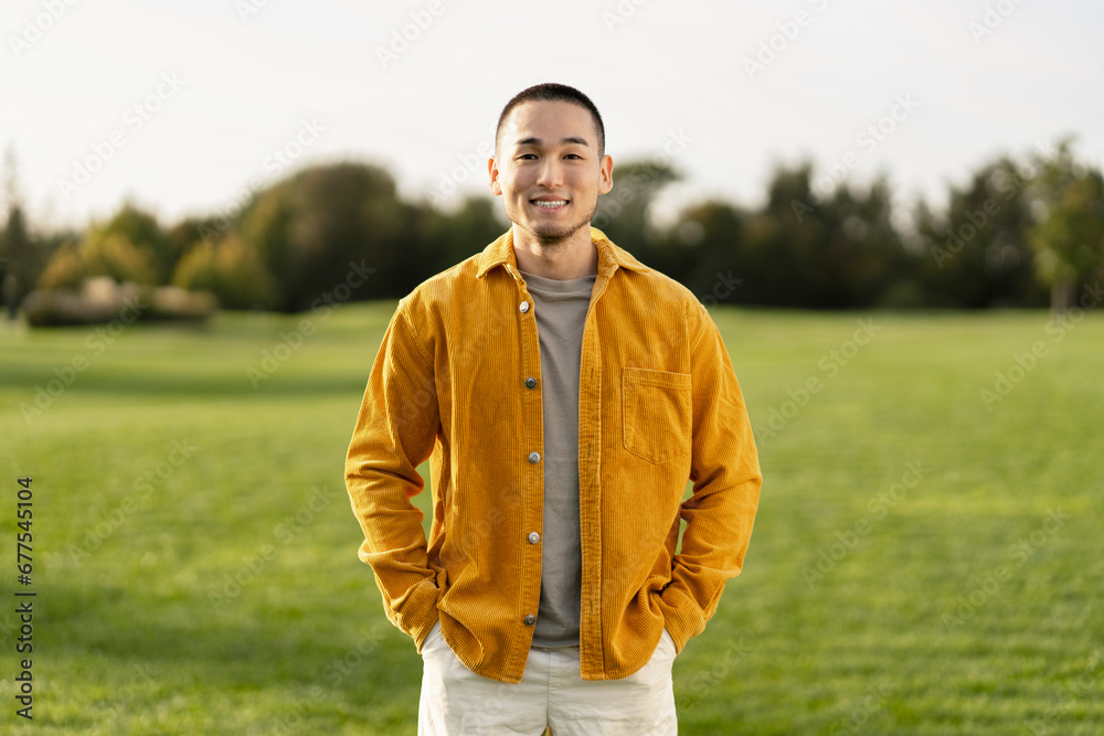Portrait of attractive smiling Asian man wearing stylish casual clothes holding hands in pockets looking at camera in green park Portrait of attractive smiling Asian man wearing stylish casual clothes holding hands in pockets looking at camera in green park