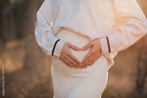 Close up tummy of pregnant woman making heart shape with hands wearing knitted sweater in sun light outdoor over nature background. Maternity lifestyle. Winter season. photo