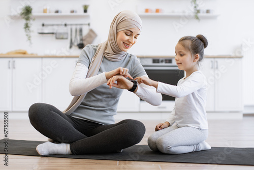 Muslim female with daughter checking workout achievements on sports watch while sitting on yoga mat in kitchen. Mother and her girl taking advantages of home training using fitness tracker.