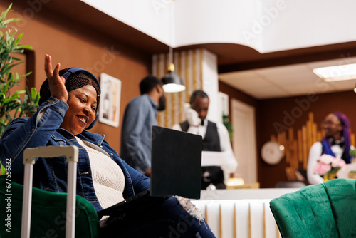 Happy smiling African American woman traveler sitting in hotel lobby having video call while waiting for check-in. Female tourist wearing headphones waving hand greeting friend looking at webcam