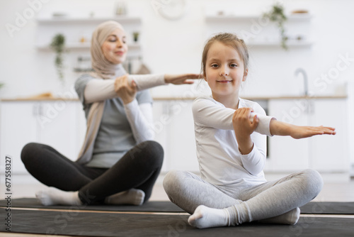 Front view of small female kid on yoga mat stretching arms with slim muslim lady with closed eyes during training. Young parent and daughter in sportswear spend time happily during workout.