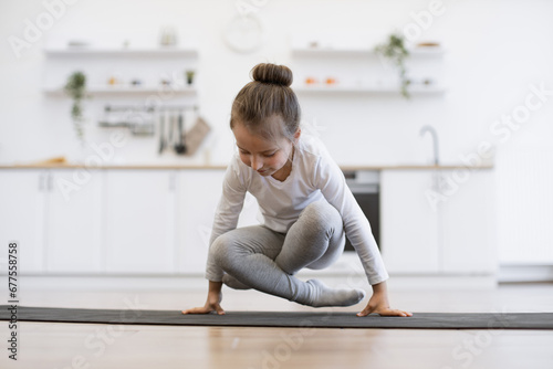 Front view of cute girl practicing yoga, standing in crane exercise, bakasana pose, working out on mat wearing sportswear, indoor full length, in white loft kitchen background.