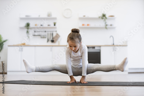 Preschool cute little girl practicing yoga  standing in crane exercise  bakasana pose  working out on mat wearing sportswear  indoor full length  in white loft kitchen background.