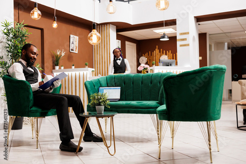 Young African American smiling man bellboy wearing uniform and white gloves sitting at hotel lounge area holding clipboard making notes. Bellhop filling information of luggage in arrival errand card