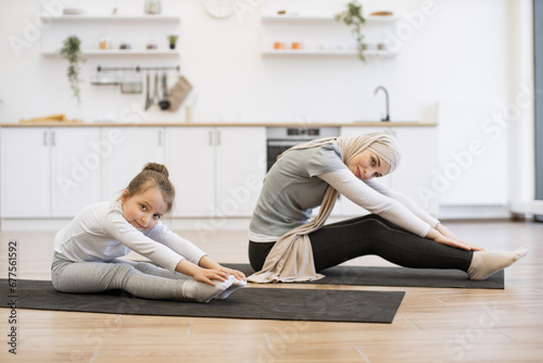 We love fun sport. Charming sportive slim active energetic beautiful muslim mum and cute lovely adorable daughter wearing sport clothes sitting on the floor mat touching toes on the outstretched legs