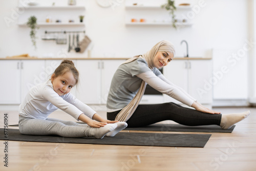 Young muslim mother in hijab showing her little daughter legs stretching exercises. Happy girls training at home. Motherhood and sporty lifestyle concept.