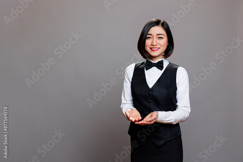 Smiling asian professional waitress showing with hands while looking at camera. Young attractive restaurant receptionist wearing black and white uniform suit and posing for studio portrait