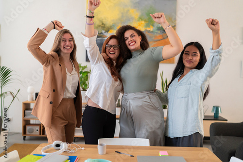 Happy young women colleagues raising arms and celebrating success while standing against blurred background in modern creative office and looking at camera photo
