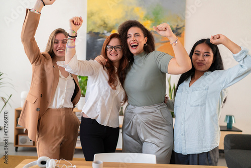 Happy young women colleagues raising arms and celebrating success while standing against blurred background in modern creative office and looking at camera photo