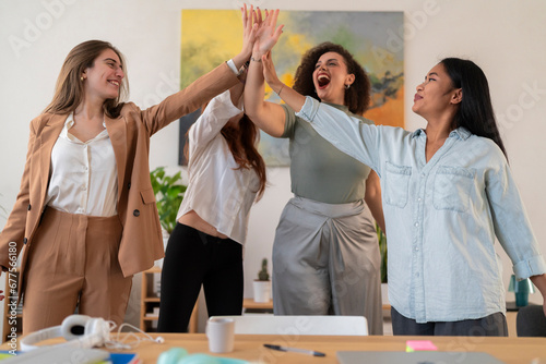 Happy young women colleagues standing and giving high five to each other while celebrating success in modern creative office during business meeting photo
