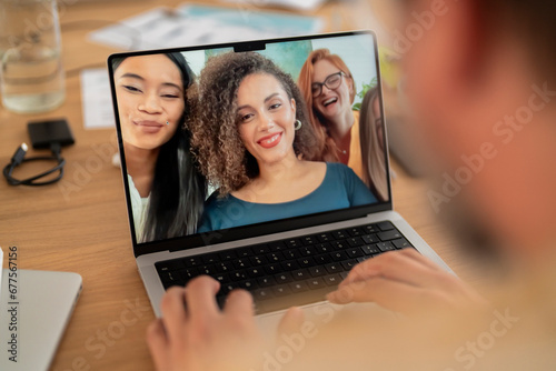 Laptop screen image of group of happy multiracial women gathered at table and taking selfie while looking at computer and working together in modern workspace photo