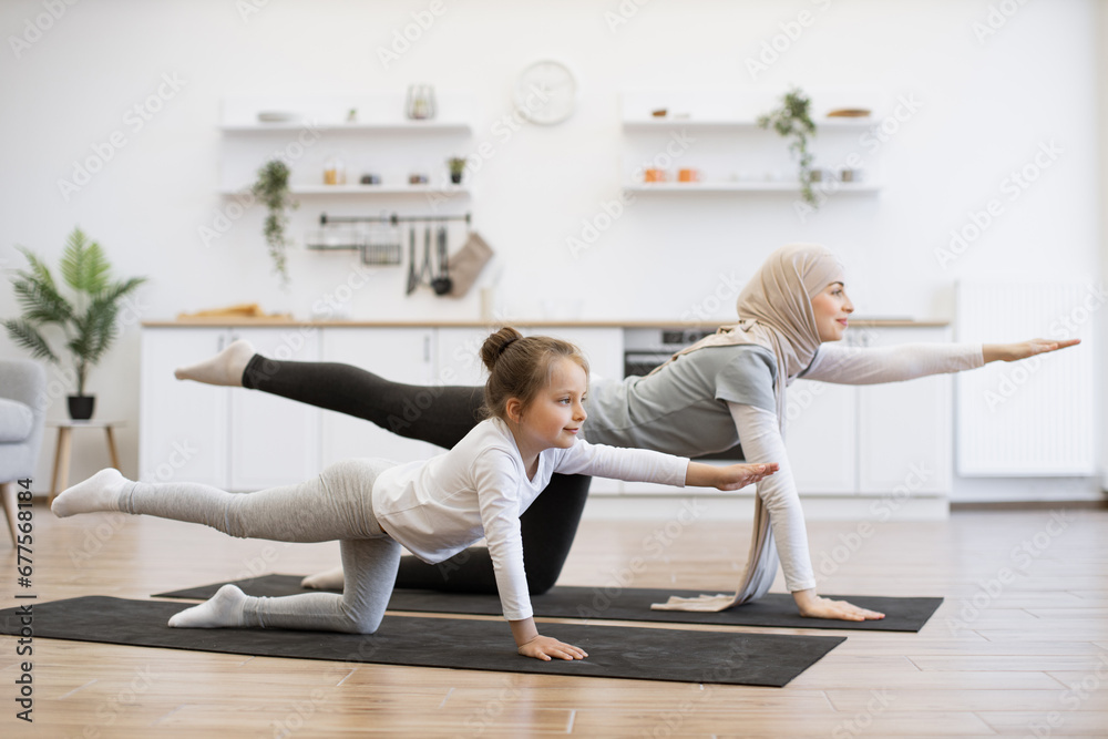 Active slim muslim lady and her daughter in sports clothes streching in balancing table pose on mats at home. Beautiful mother with her girl practicing yoga exercise together in kitchen. Active slim muslim lady and her daughter in sports clothes streching in balancing table pose on mats at home. Beautiful mother with her girl practicing yoga exercise together in kitchen.