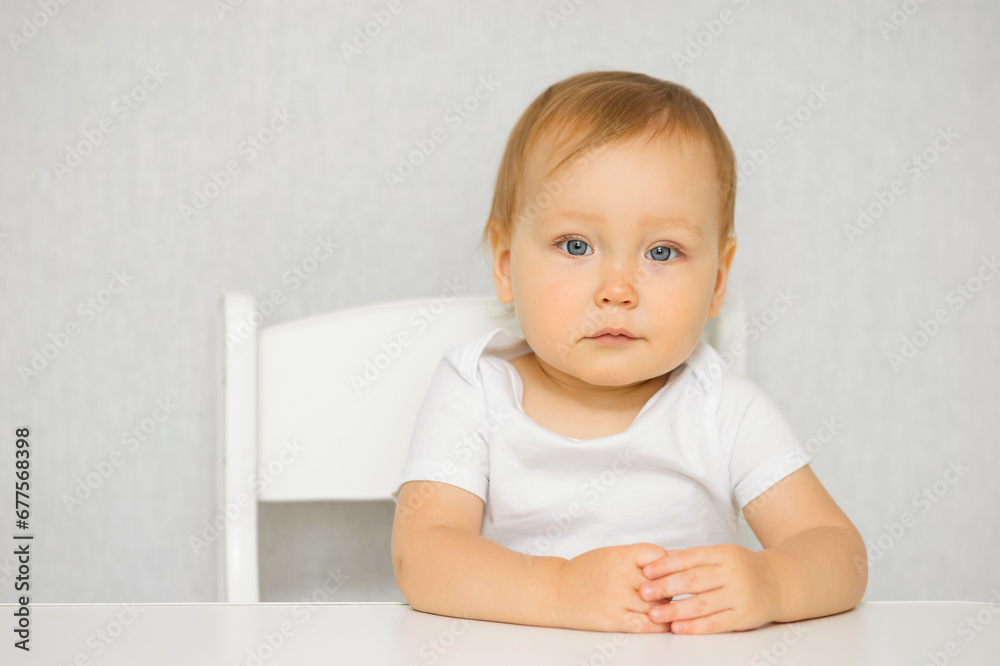 Cute little girl in T shirt sits calmly at table on chair with folded hands. Toddler blue eyed