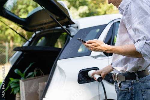 Man with smartphone next to a charging electric car in the yard of a country house