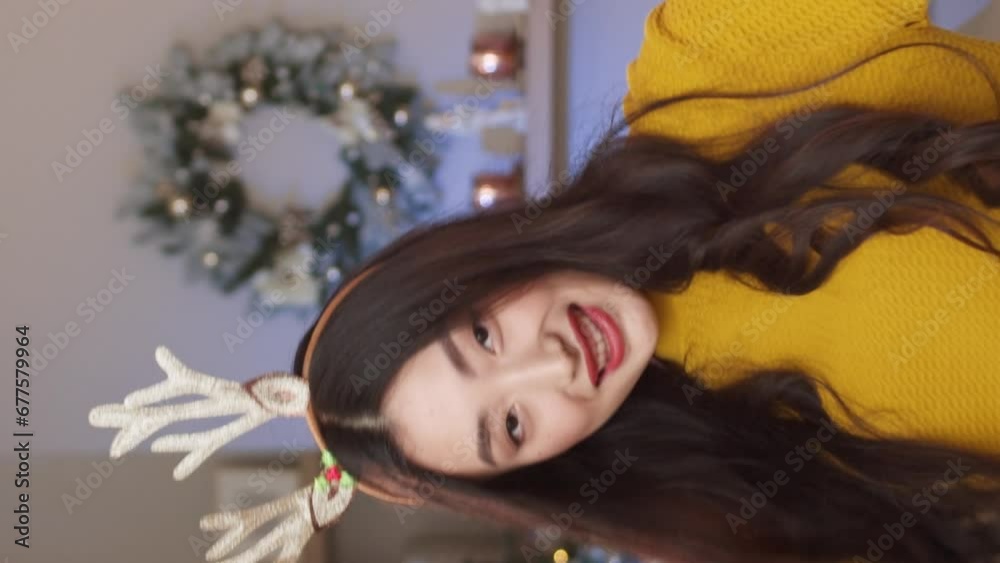 Vertical orientation of Asian woman wearing hoop with deer antlers while celebrating holiday in cozy room with Christmas tree and decorations. Young girl with bengal fire positively dancing. Vertical orientation of Asian woman wearing hoop with deer antlers while celebrating holiday in cozy room with Christmas tree and decorations. Young girl with bengal fire positively dancing.