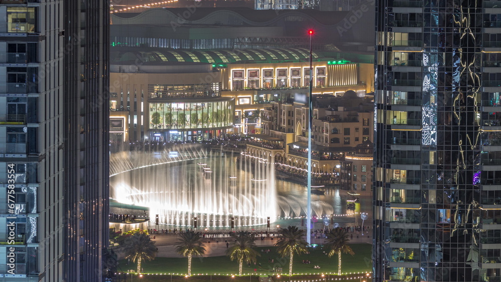 Aerial view of Dubai Fountain in downtown with palms in park next to shopping mall and souq night timelapse, UAE