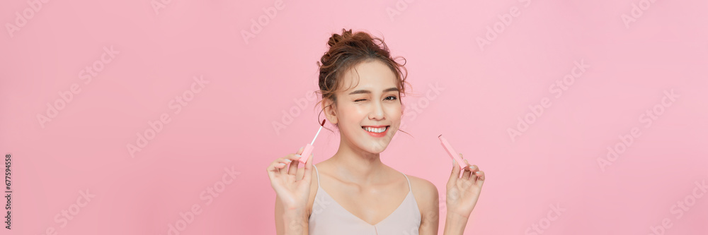 Young woman using lip gloss looking at camera on pink studio background - obrazy, fototapety, plakaty 