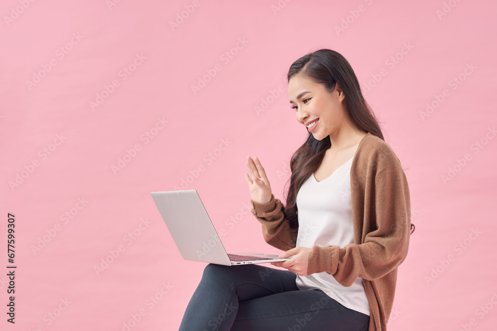 Happy young woman working online, sitting on chair and using laptop against pink wall