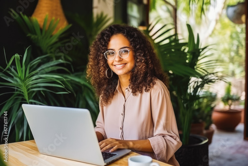 Smiling young woman with glasses using laptop at a cafe surrounded by plants. Studying for class. Medicine student.