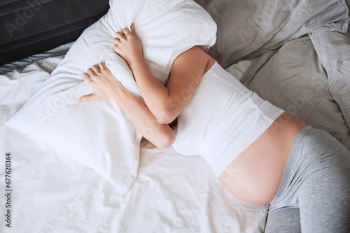 Exhausted pregnant woman suffering from insomnia, pain lying in bed covered with pillow. photo